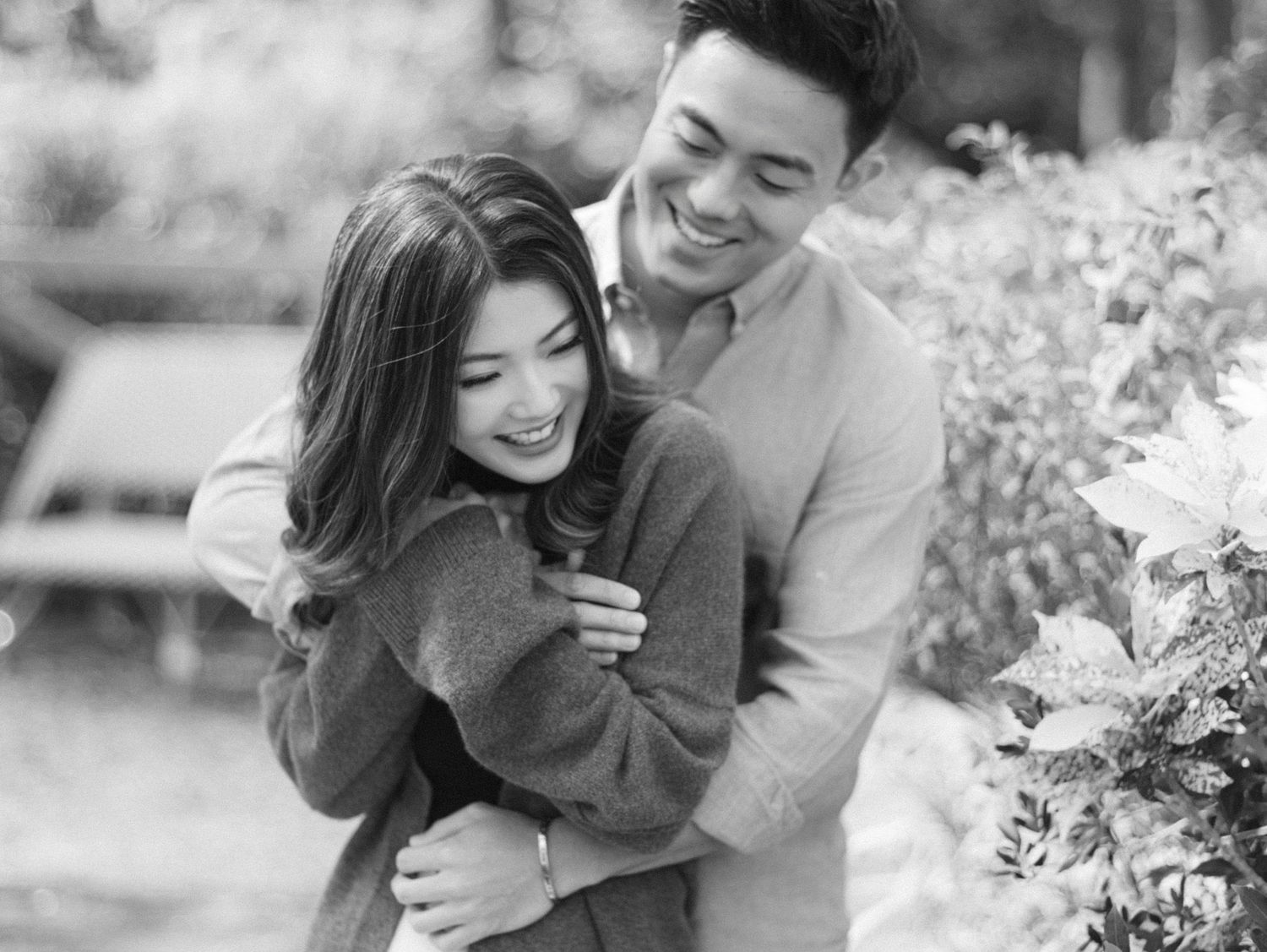 A happy Asian couple embraces and laughs together outdoors in a black and white engagement photo.
