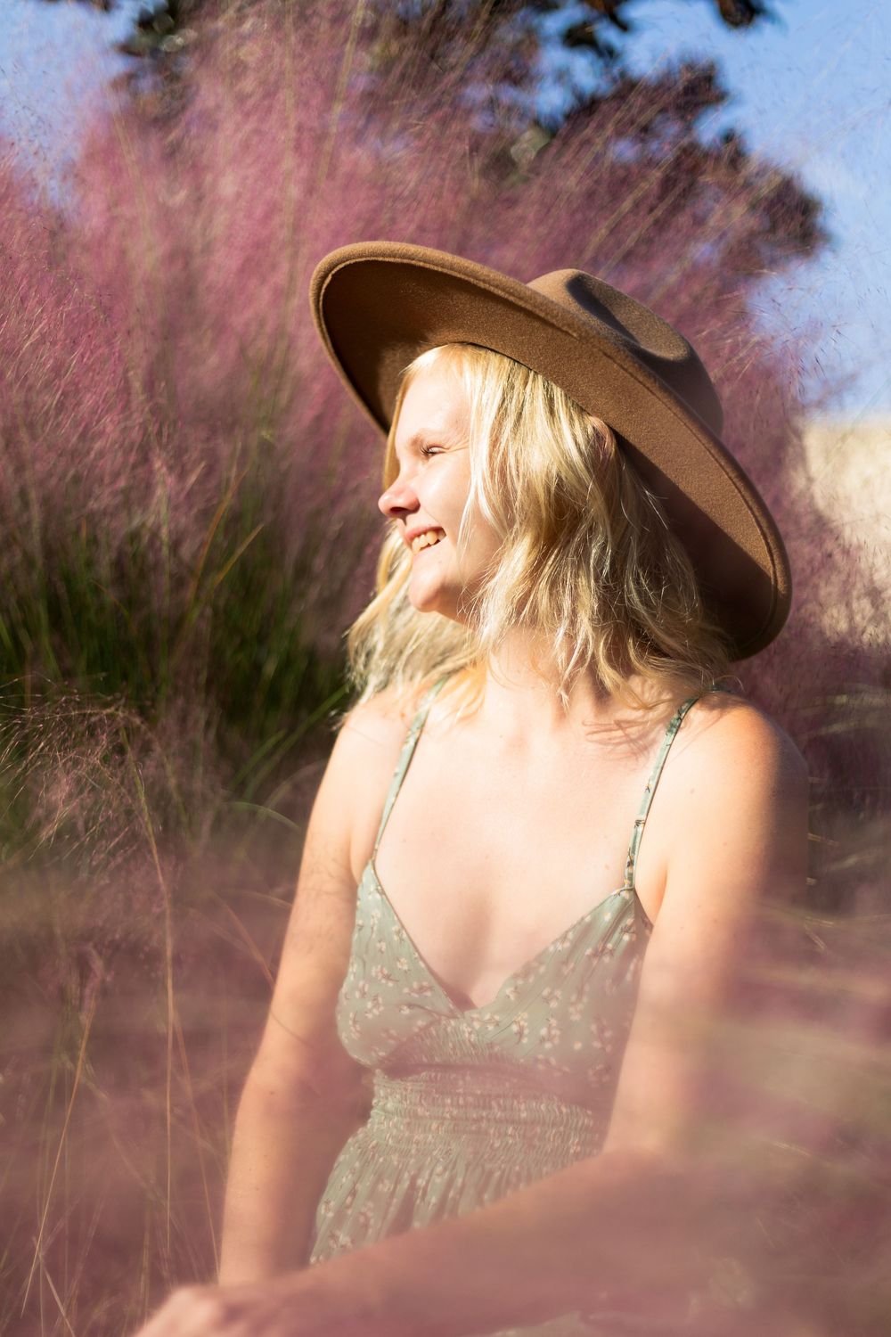 A person in a wide-brimmed hat and dress stands among pink muhly grass swaying in warm sunlight.