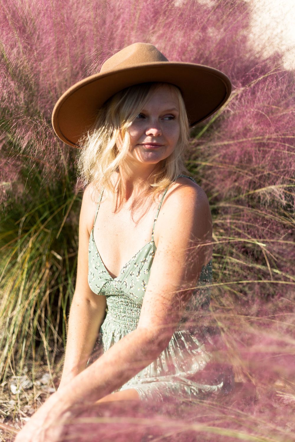 A person in a brown wide-brimmed hat and mint green dress poses among pink muhly grass in soft sunlight.
