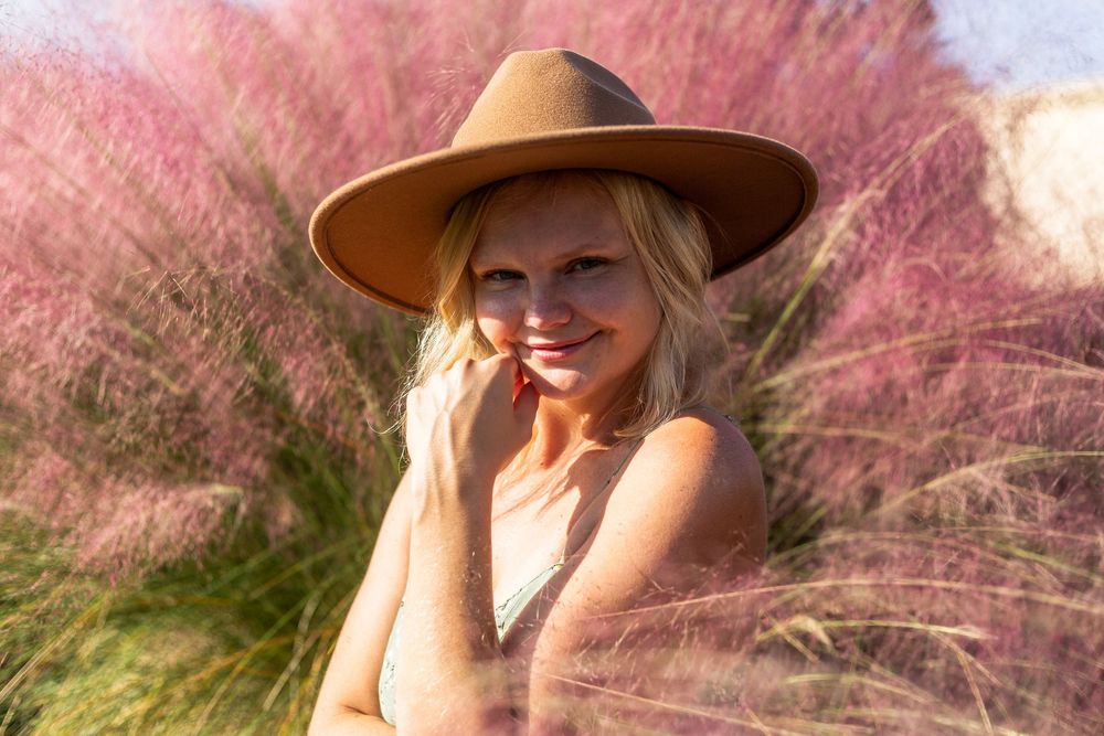 Someone wearing a brown hat smiles outdoors in front of pink ornamental grass on a sunny day.