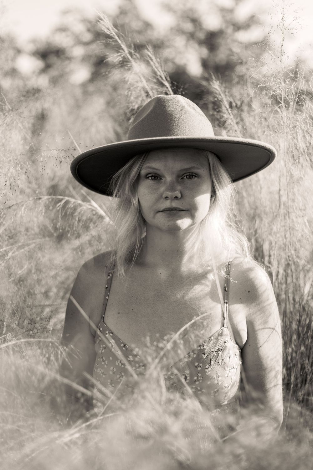 A black and white portrait shows a figure in a wide-brimmed hat amidst tall wild grasses.