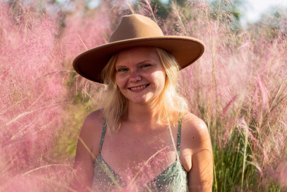 Someone wearing a brown hat and floral dress poses in a field of pink muhly grass on a sunny day.