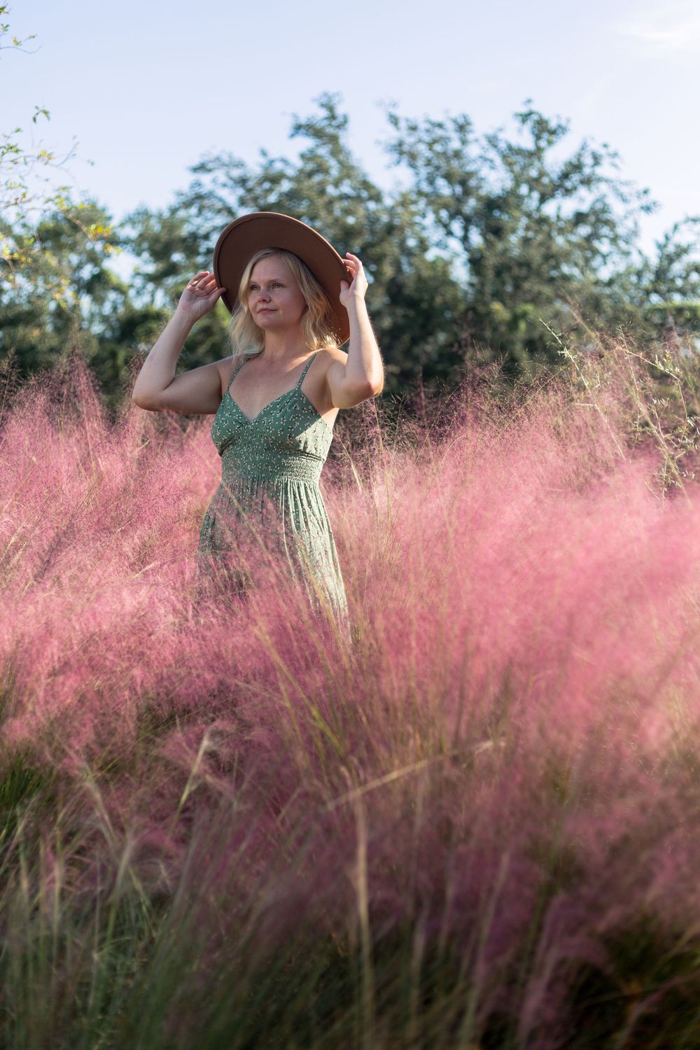 Person in a green dress and wide-brimmed hat poses elegantly among pink muhly grass in a dreamy outdoor setting.