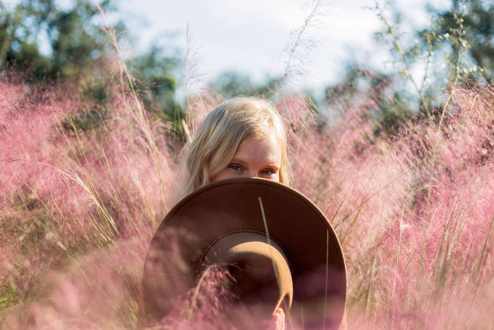 Person holding a camera lens surrounded by pink wispy grass flowers in a dreamy outdoor setting.