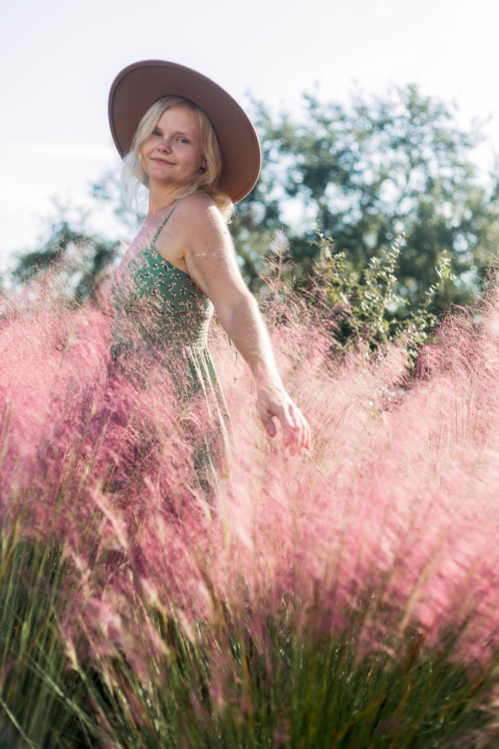 Person wearing a hat smiles while standing in a field of flowing pink muhly grass during golden hour.