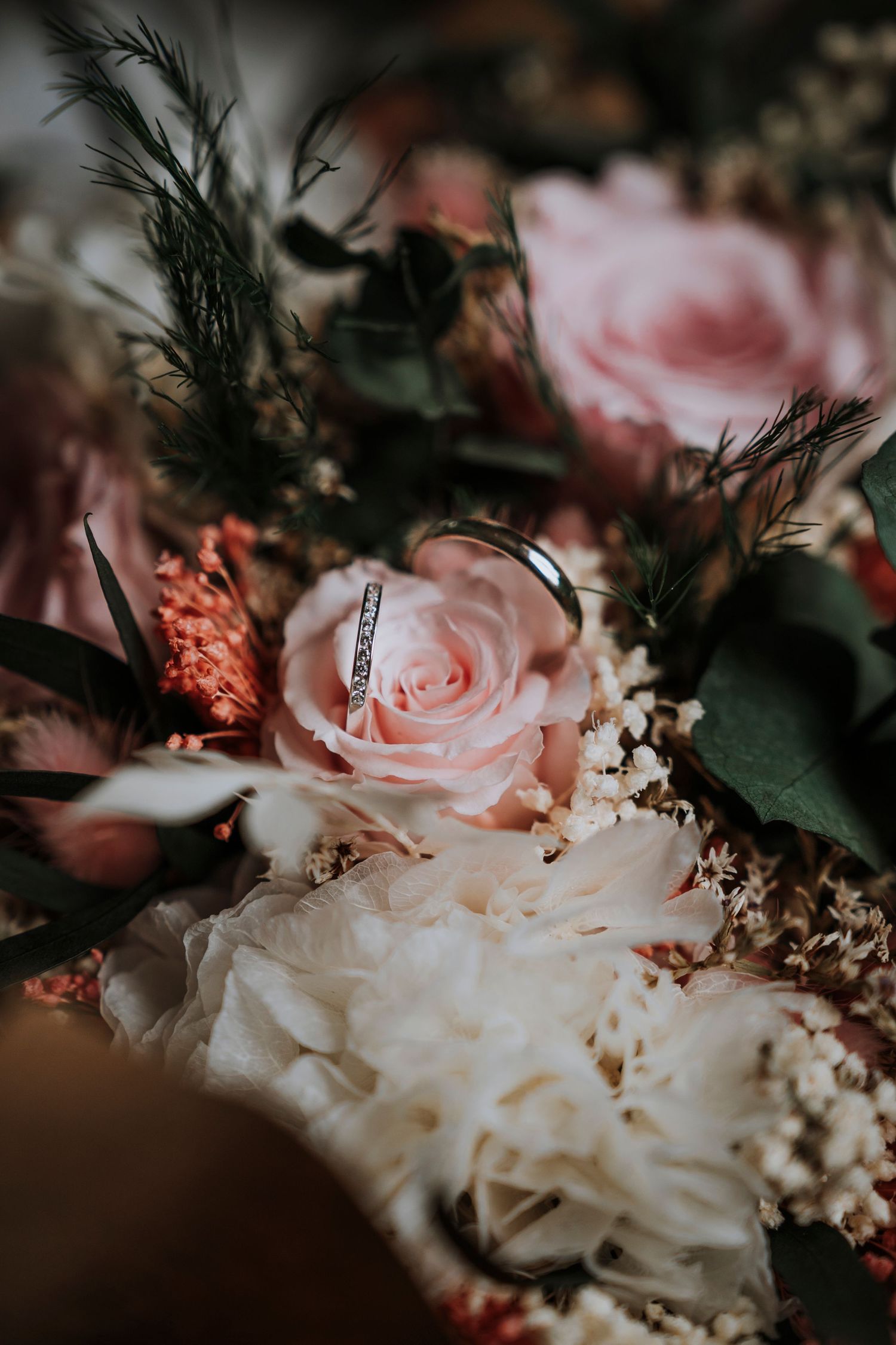 A diamond wedding ring resting on a pink rose in a romantic bridal bouquet with greenery and dried flowers.