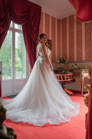 An elegant wedding dress with flowing tulle train photographed in a room with red curtains and carpet.
