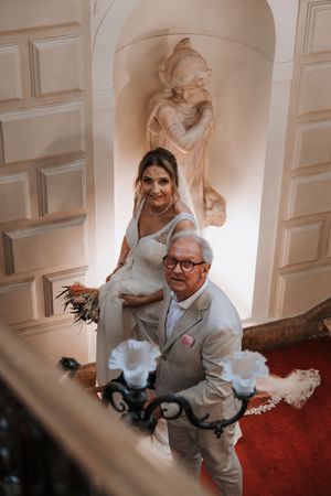 Bride in white gown and older man in gray suit ascending elegant staircase with classical statue backdrop.