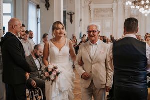 Bride walks down the aisle with older gentleman in beige suit, holding pink bouquet in elegant ceremony hall.