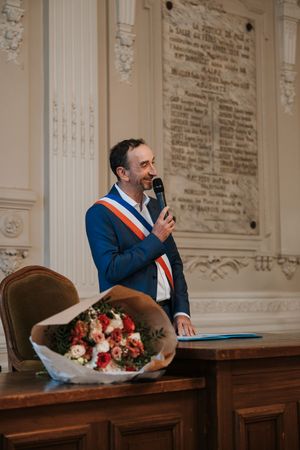 Man in blue suit with French mayoral sash speaking into microphone at a wedding ceremony.