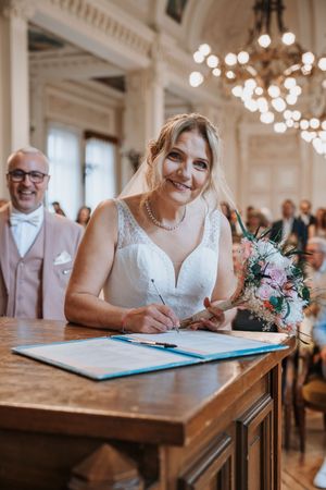 Smiling bride signing wedding register at ceremony venue holding pink bouquet with groom behind.