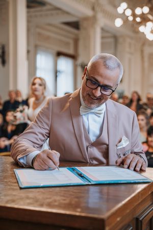 A groom in a beige suit and bow tie signing the wedding register at a ceremony venue.
