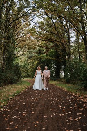 Bride and groom walking hand-in-hand down a tree-lined autumn path on their wedding day.