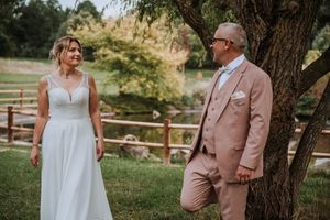 Bride in white gown and groom in pink suit share a romantic moment outdoors near a tree by a scenic pond.