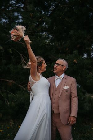 Bride in white gown raises bouquet while groom in pink suit smiles outdoors near lush green trees.