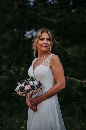 Bride in white lace gown holding floral bouquet outdoors.