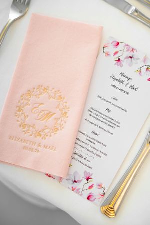 Elegant pink wedding menu and napkin with gold monogram and floral design on white place setting.