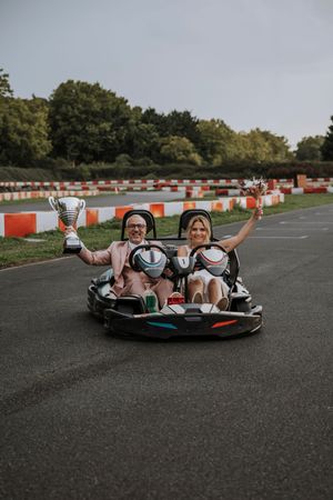 Couple celebrating in a go-kart holding trophy cups on a race track with orange barriers.