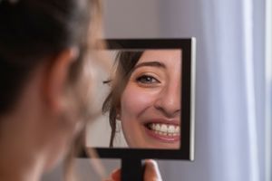 Young woman smiling at her reflection in a handheld mirror, showcasing bright white teeth and natural beauty.