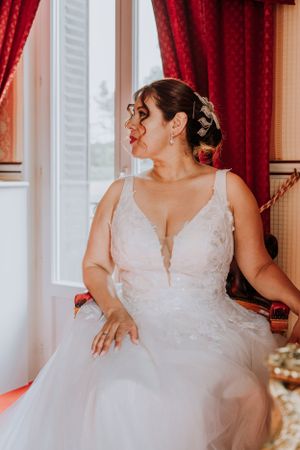 A white wedding dress with tulle skirt and lace bodice displayed in an elegant indoor setting with red curtains.