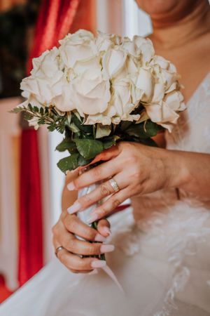 Close-up of hands holding a white rose bouquet, showing wedding rings and lace dress details.