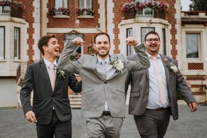 Groomsmen in grey suits and dark ties laughing together outside a brick building.