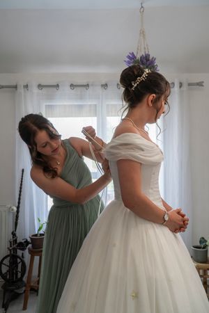 Bridesmaid in green dress helping bride with white ball gown wedding dress in bright room.