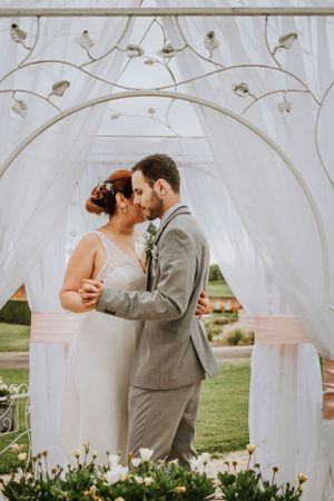 A romantic moment under a white draped wedding arch in an outdoor garden setting.