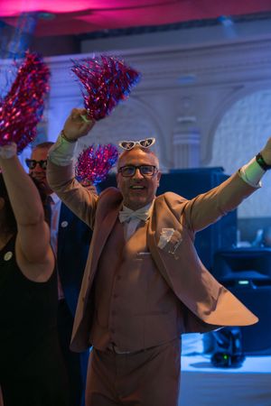 Joyful man in tan suit celebrating at wedding reception with red pom-poms and sunglasses on his head.