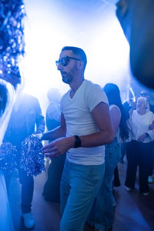 Man in white t-shirt and sunglasses dancing at a nightclub party with blue lighting.