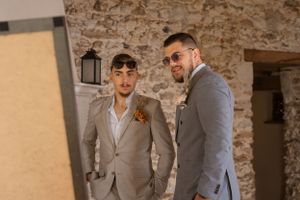 Groomsmen in light grey suits gather in rustic stone-walled venue.