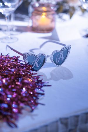 Heart-shaped wedding sunglasses with initials on white table beside pink tinsel and glowing candles.