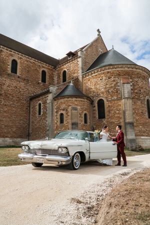 Bride and groom pose beside a vintage white classic car in front of a historic stone church.