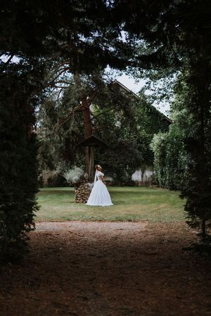 Bride in white gown standing in lush garden framed by tall trees near rustic building.