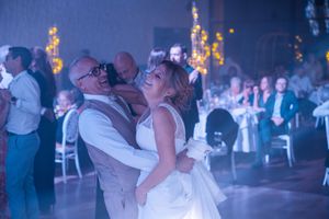 Bride and groom share joyful first dance embrace on lit reception dance floor with guests watching.
