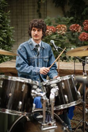 Young male drummer playing drums outdoors wearing denim jacket with drumsticks at garden event.