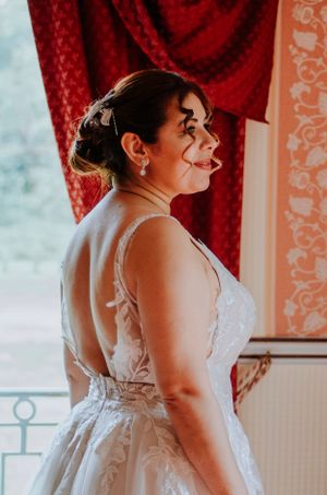 Side profile of someone in a white lace wedding dress near a window with red curtains.