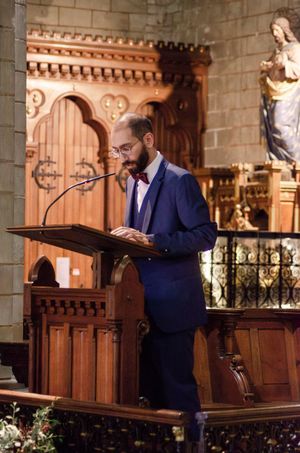Man in blue suit speaking at a wooden church pulpit with religious statues in the background.