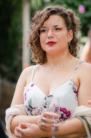 Woman in floral dress holding wine glass wearing glasses and gold earrings at outdoor event.