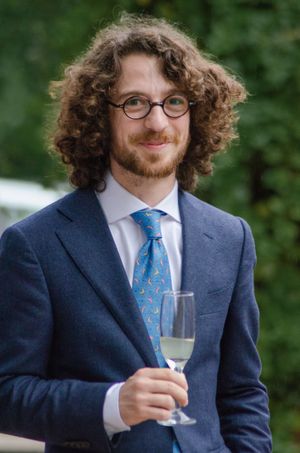 Young man with curly hair and glasses holding a champagne glass at an outdoor wedding reception.