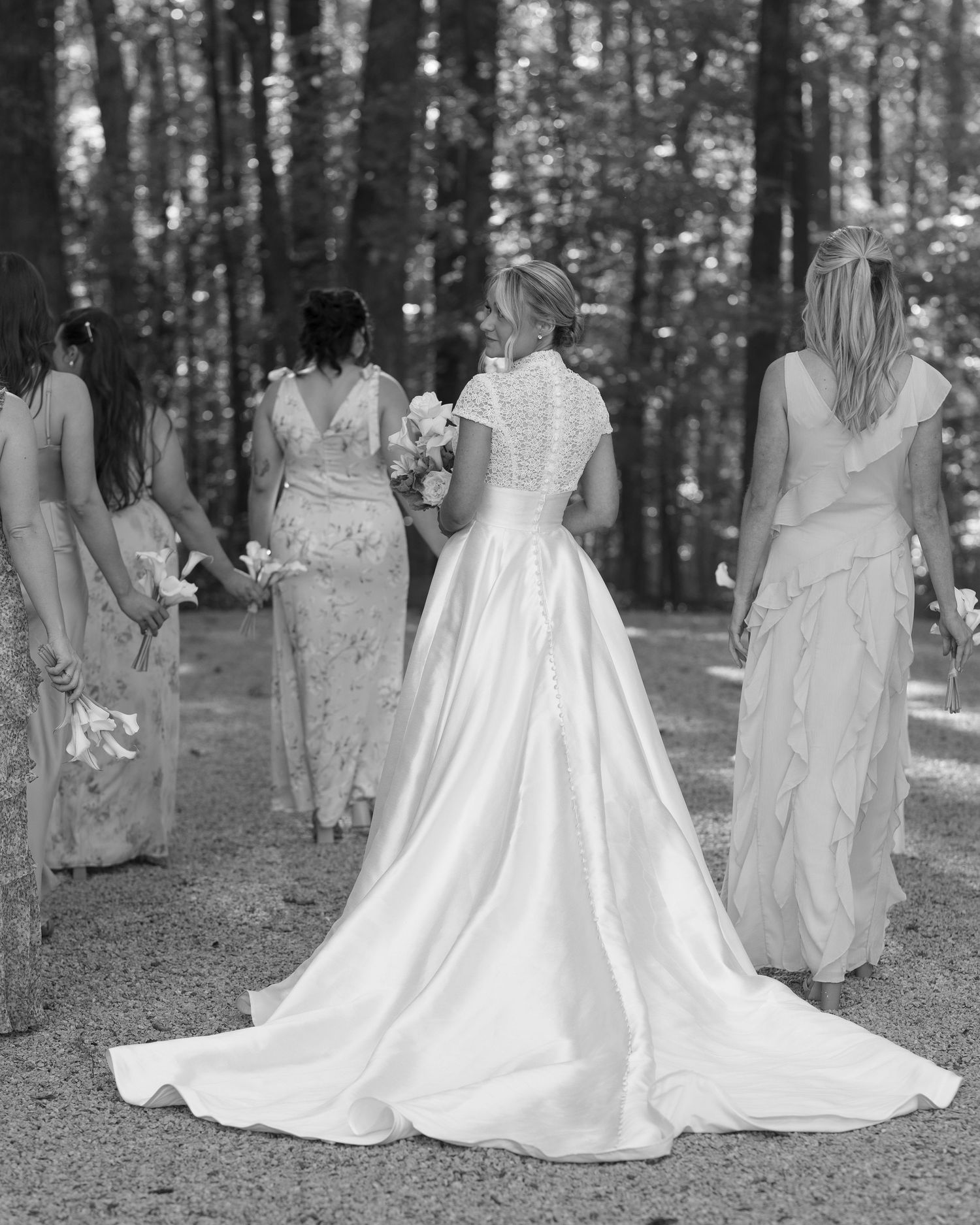 Wedding party wearing elegant white dresses stands together in a wooded outdoor setting.