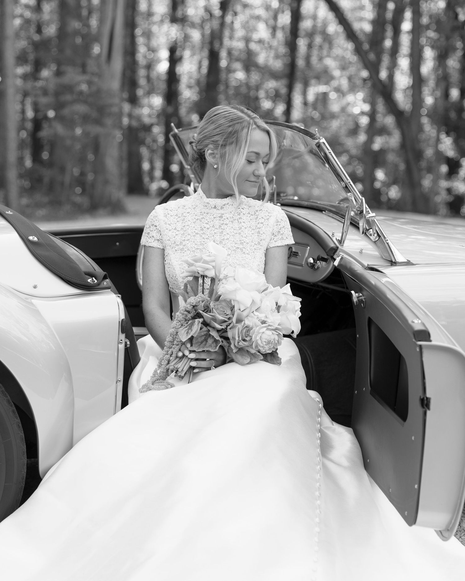 Row of white wedding dresses lined up against vintage cars in black and white photography.