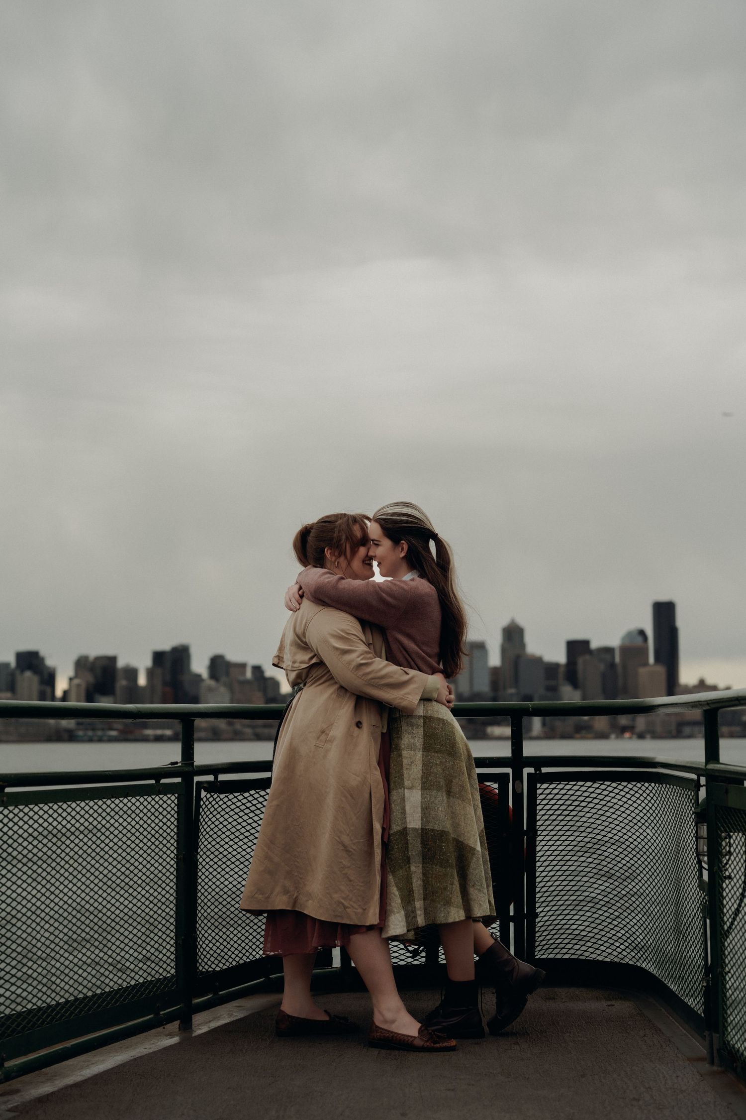 A couple shares an intimate moment on a ferry deck with a city skyline in the background.