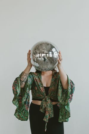 Person wearing green floral kimono top and black pants holding disco ball in front of their face in artistic portrait.