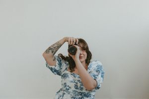 Someone wearing a floral print dress holds an analog camera up to their face while posing against a white wall.