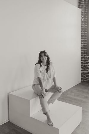 Black and white portrait of a person in a white shirt sitting casually on a white step against a brick wall backdrop.