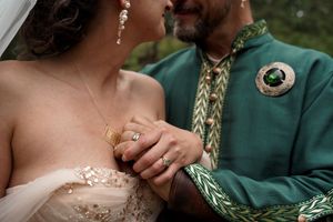 Close-up detail of wedding rings and formal attire showing emerald green jacket with gold embroidery and pearl beaded dress.