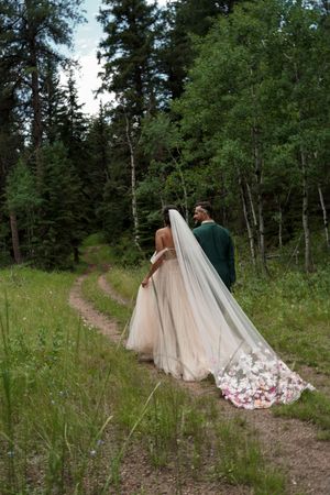 A flowing blush wedding dress trails behind on a dirt path through a green forest setting.