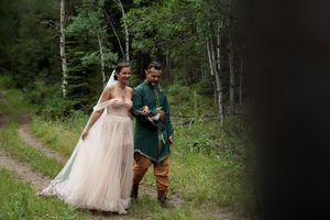 A romantic couple in wedding attire walks hand in hand along a wooded forest path.