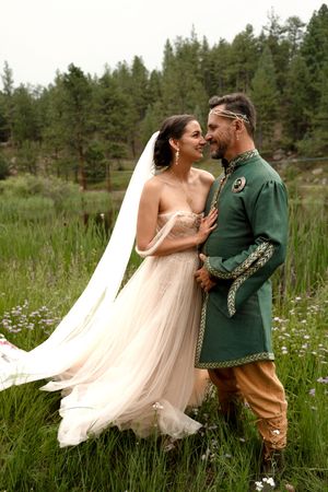 A couple in fairy tale wedding attire embracing in a meadow with pine trees in the background.